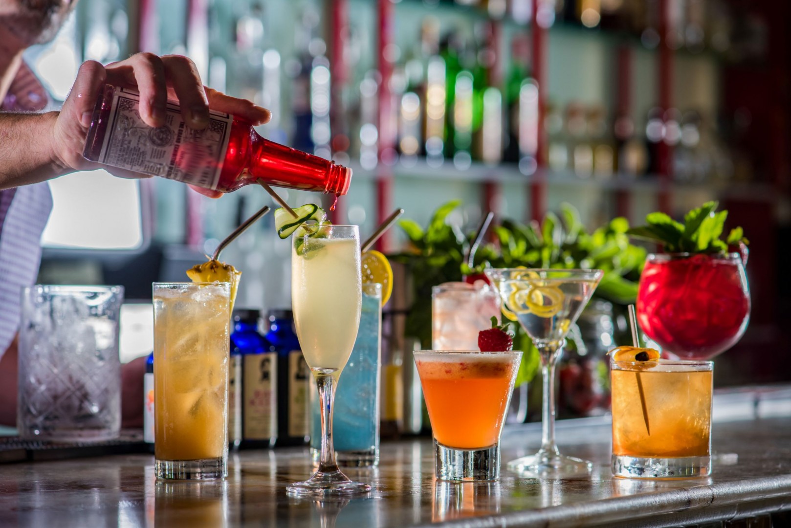bartender's hand pouring beverage into a cocktail glass, surrounded by other cocktails of different colors and glass styles